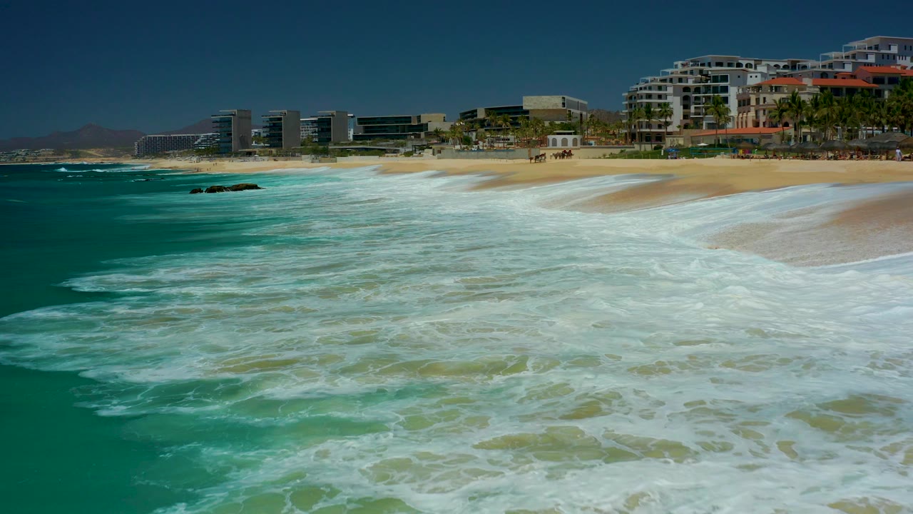 At 0:03, churning white surf covers more of the lower right foreground, with turquoise water to the left and the Marquis Los Cabos beach buildings across the top of frame.