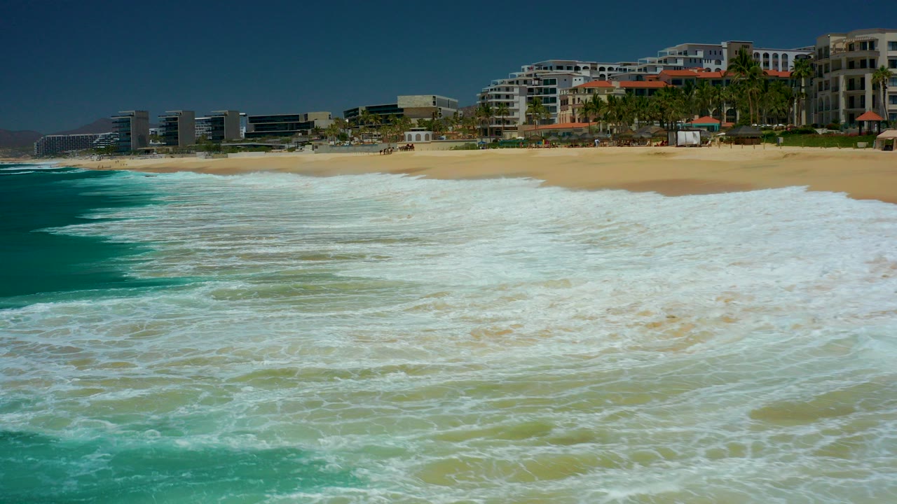 At 0:00, white surf fills the foreground as waves approach the sandy beach at Marquis Los Cabos, with resort buildings, palms, distant hills, blue sky, and no visible people.