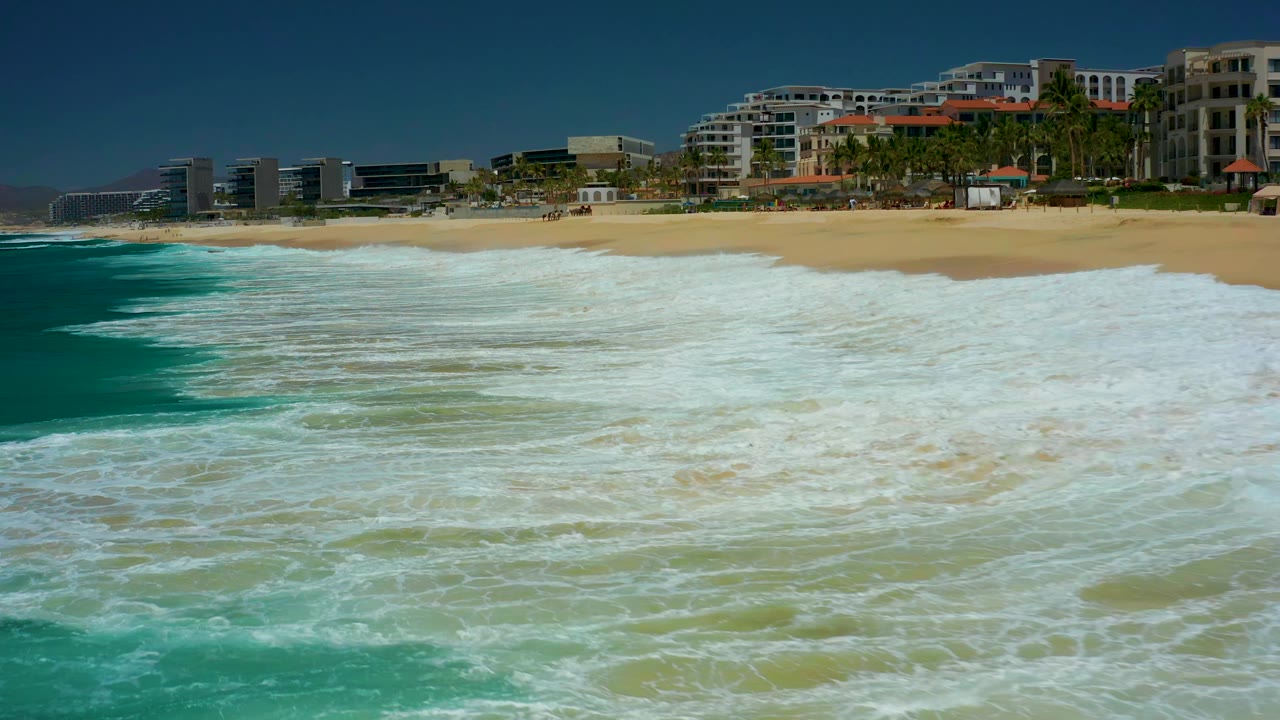 At 0:01, the foam advances across shallow turquoise water toward the sand at Marquis Los Cabos, with modern coastal buildings and palms lining the beach, and no visible people.