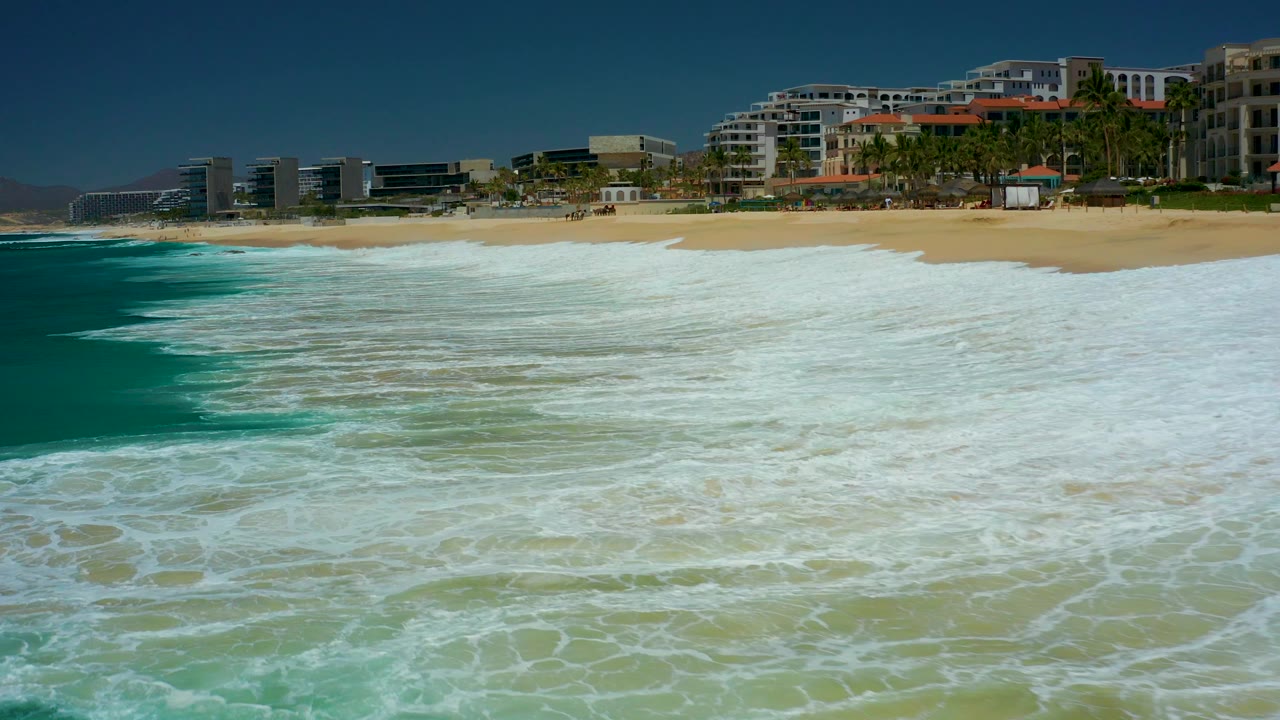 At 0:02, a broad band of white water reaches the beach edge at Marquis Los Cabos, while the background buildings, palms, distant hills, and clear sky remain visible, with no visible people.