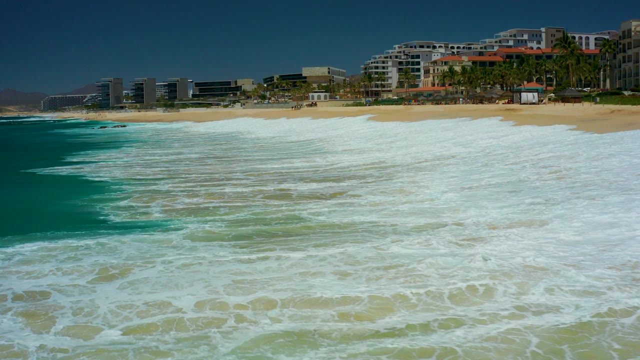 At 0:03, foamy ocean water covers most of the foreground at Marquis Los Cabos, with a sandy strip, beachfront buildings, palms, low mountains, and no visible people in view.