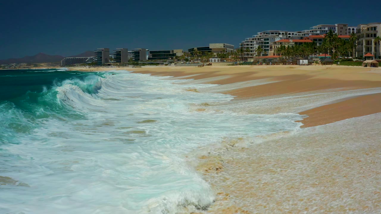 White foam advances diagonally across the Marquis Los Cabos shoreline, with deep blue water on the left, tan beach on the right, background buildings, palms, and no visible people.