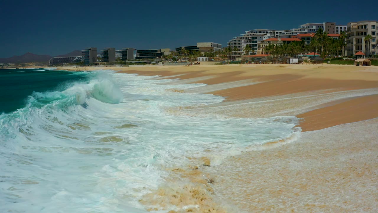 A larger crest curls and collapses near the Marquis Los Cabos beach, sending white water across the sand below background resort buildings and palms, with no visible people.