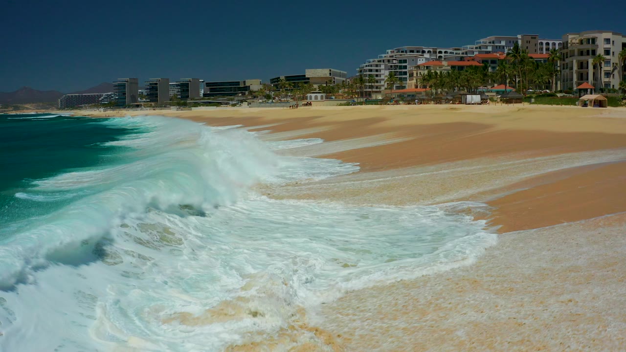 Wide white foam blankets the lower part of the Marquis Los Cabos beach while darker turquoise water remains at left and coastal buildings sit beyond the sand, with no visible people.
