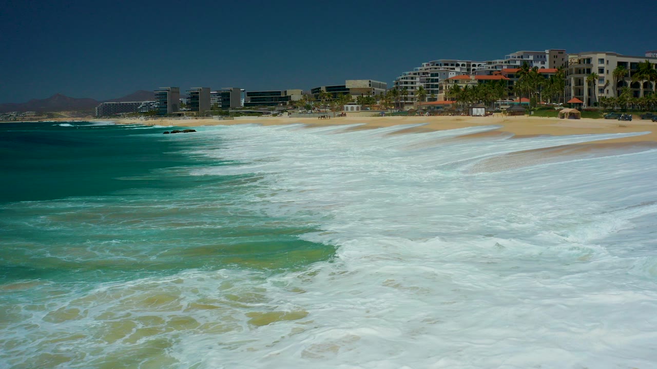 At Marquis Los Cabos in Baja California Sur, white ocean foam spreads across turquoise water toward a sandy beach with resort buildings and palms in the background, with no visible people.