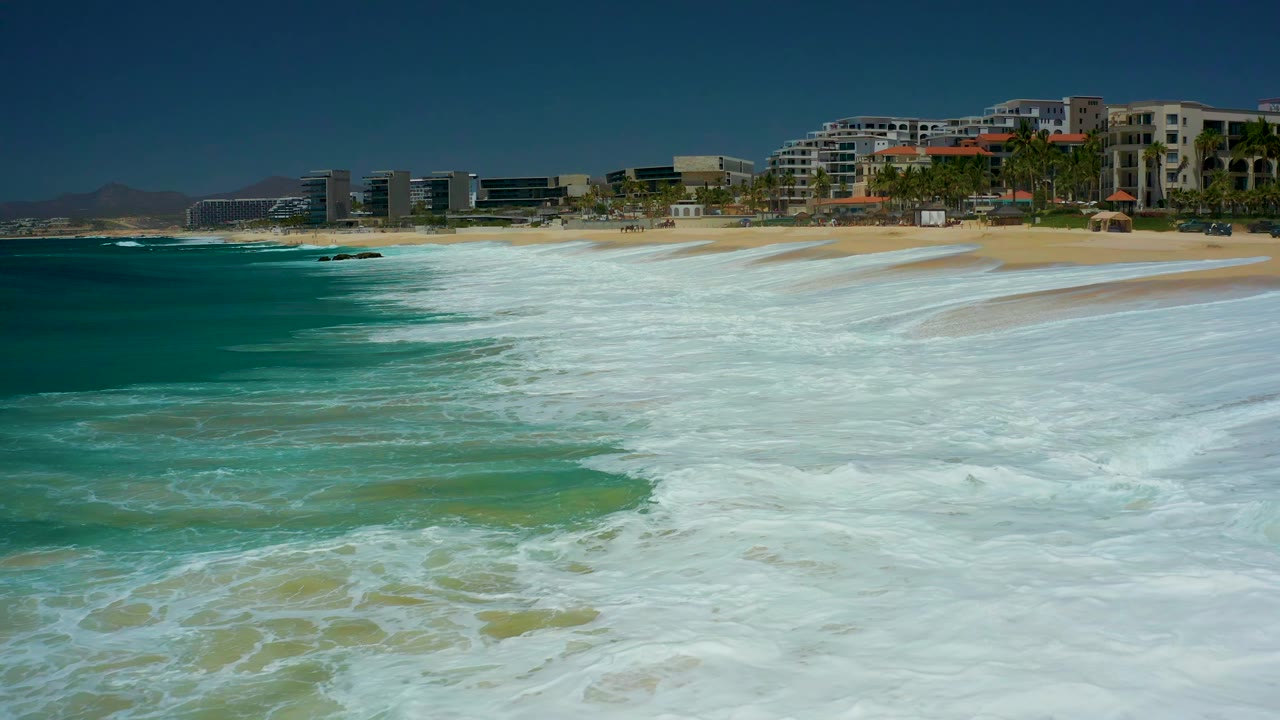 Whitewater widens across the foreground at Marquis Los Cabos while the sandy shoreline and background coastal buildings remain visible under bright daylight, with no visible people.