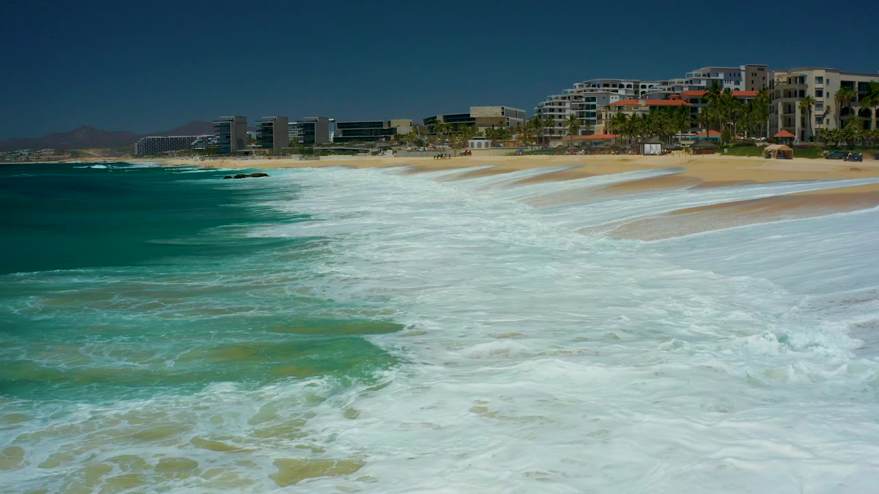 A larger sheet of white surf reaches the tan beach at Marquis Los Cabos, with turquoise water to the left and resort-style buildings lining the background, with no visible people.