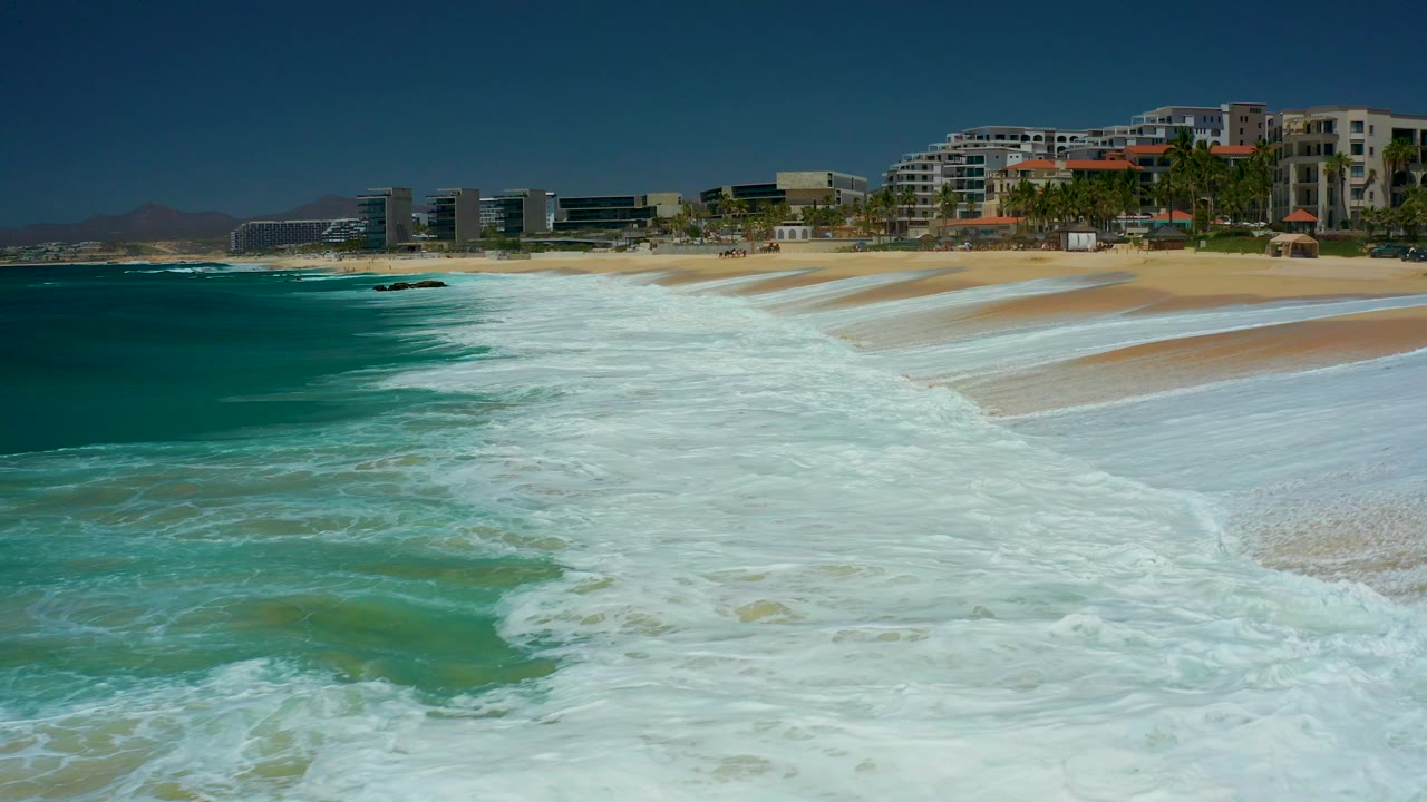 The frame shows deeper turquoise water on the left, a broad white foam path in the center, and the sandy Marquis Los Cabos shoreline backed by buildings and palms, with no visible people.