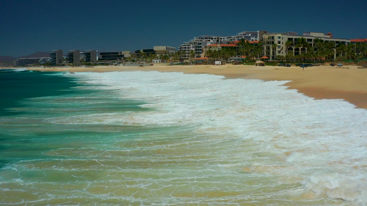 White surf spreads over turquoise water toward a sandy beach at Marquis Los Cabos, with coastal buildings and palm trees in the background and no visible people.