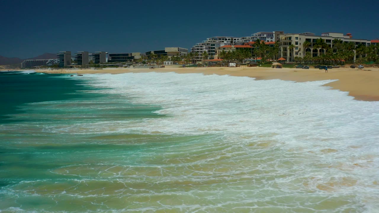 Breaking white surf pushes farther across the foreground at Marquis Los Cabos while the sandy beach, palms, and coastal buildings remain visible under clear blue sky, with no visible people.