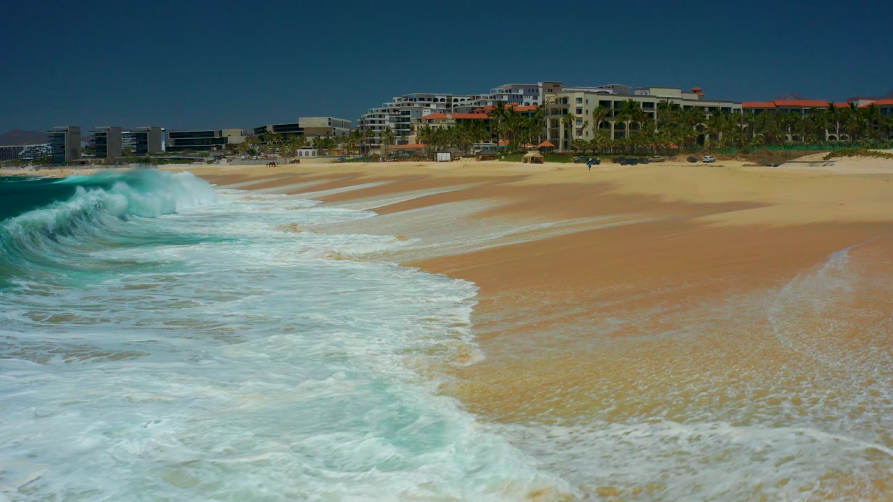 Breaking wave and white foam reaching a sandy beach with coastal buildings behind it.