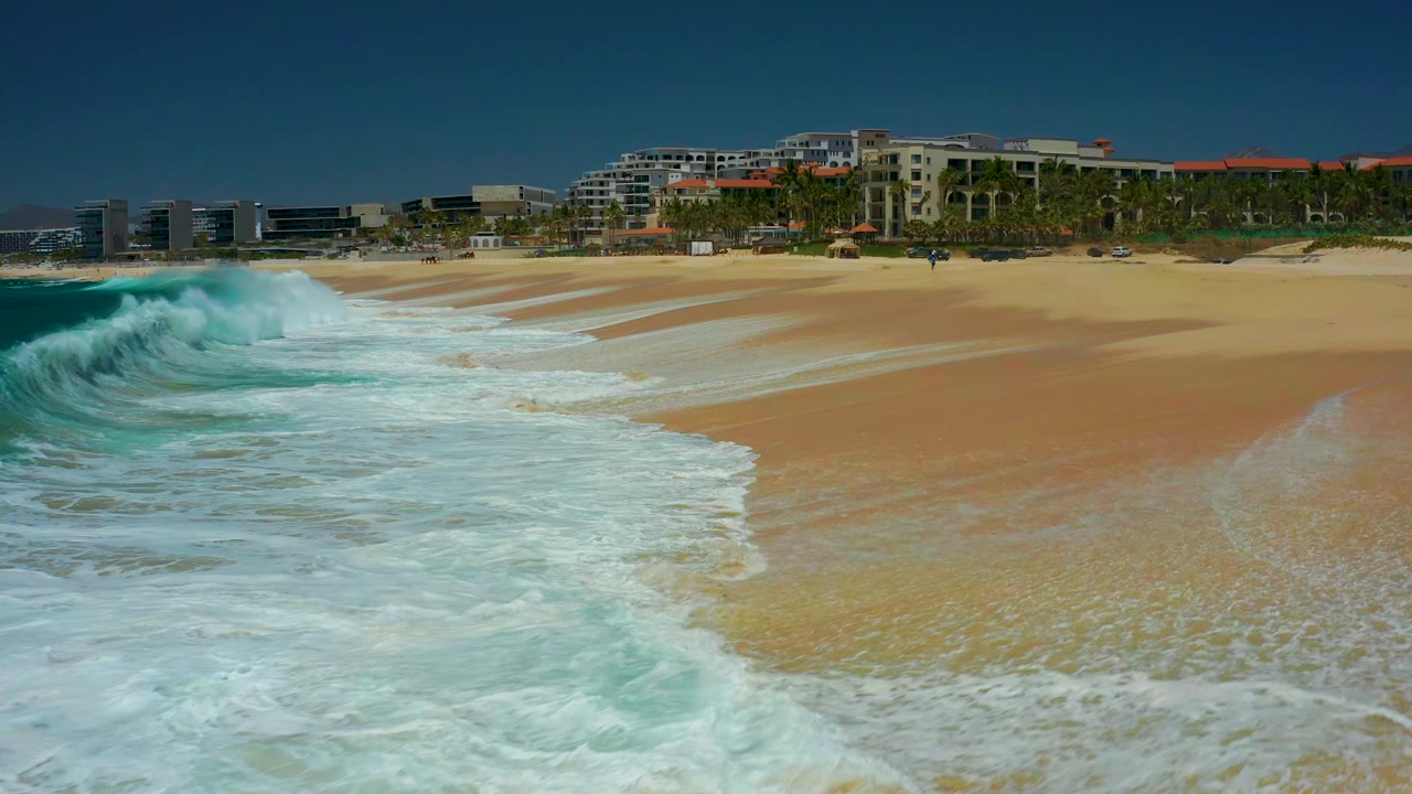 At 0:01, white water pushes farther across the Marquis Los Cabos shoreline, with wet sand bands leading toward low coastal buildings and no visible people.