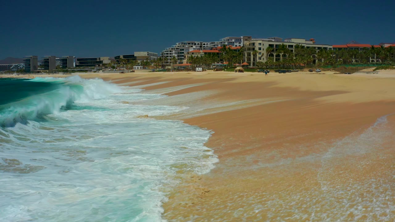 At 0:02, the foam line curves diagonally across the sand at Marquis Los Cabos, with turquoise water at left, buildings beyond the beach, and no visible people.