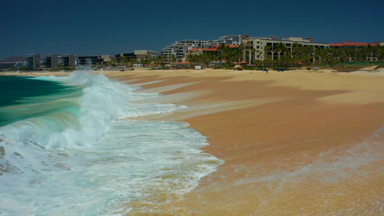 At 0:03, a larger white wave breaks in the left foreground while the empty tan beach and Marquis Los Cabos building line remain visible behind it.