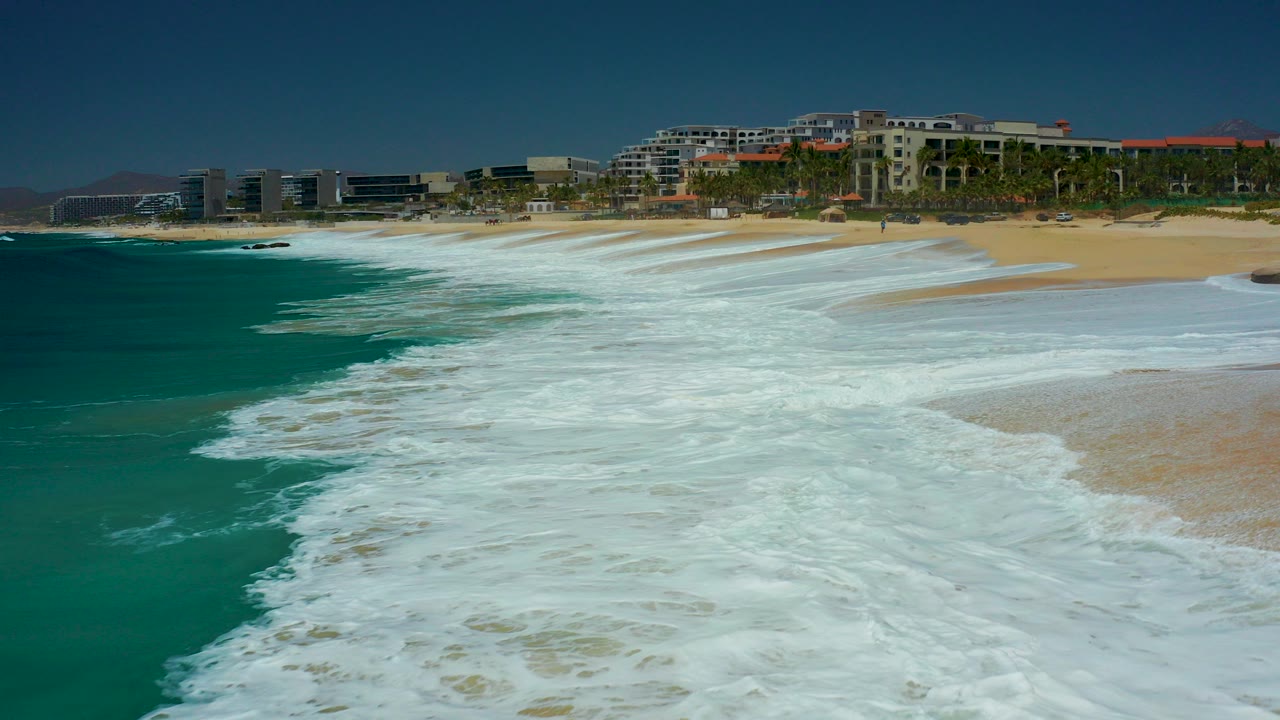 Turquoise surf spreads across a sandy beach with resort buildings and palm trees behind it.