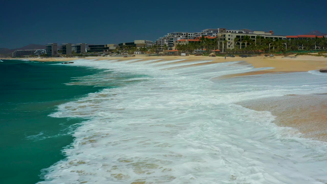 A broad sheet of white foam rolls across the foreground at Marquis Los Cabos, with tan sand, palm trees, and resort buildings visible beyond it, with no visible people.