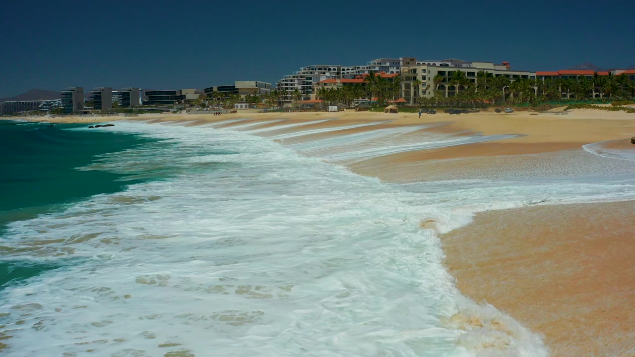 At Marquis Los Cabos, a curling wave breaks near the left edge while white foam trails over tan sand and the resort frontage remains in the distance, with no visible people.