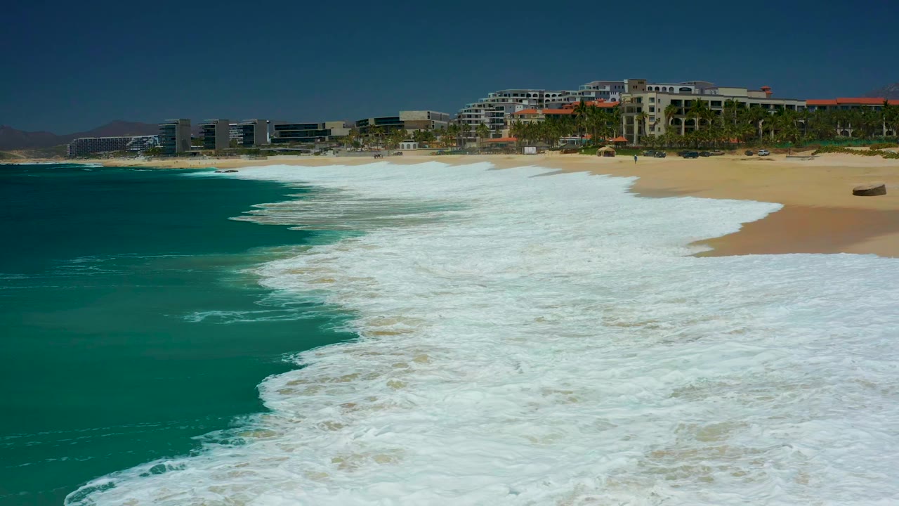 At Marquis Los Cabos, a wide band of white foam advances over the tan beach below resort-area buildings, palms, blue sky, and no visible people.