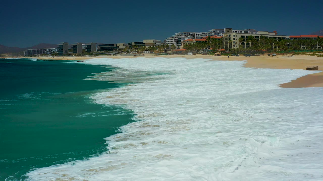 At Marquis Los Cabos, the surf forms a diagonal edge between turquoise ocean and tan sand beneath a row of coastal buildings and palms, with no visible people.