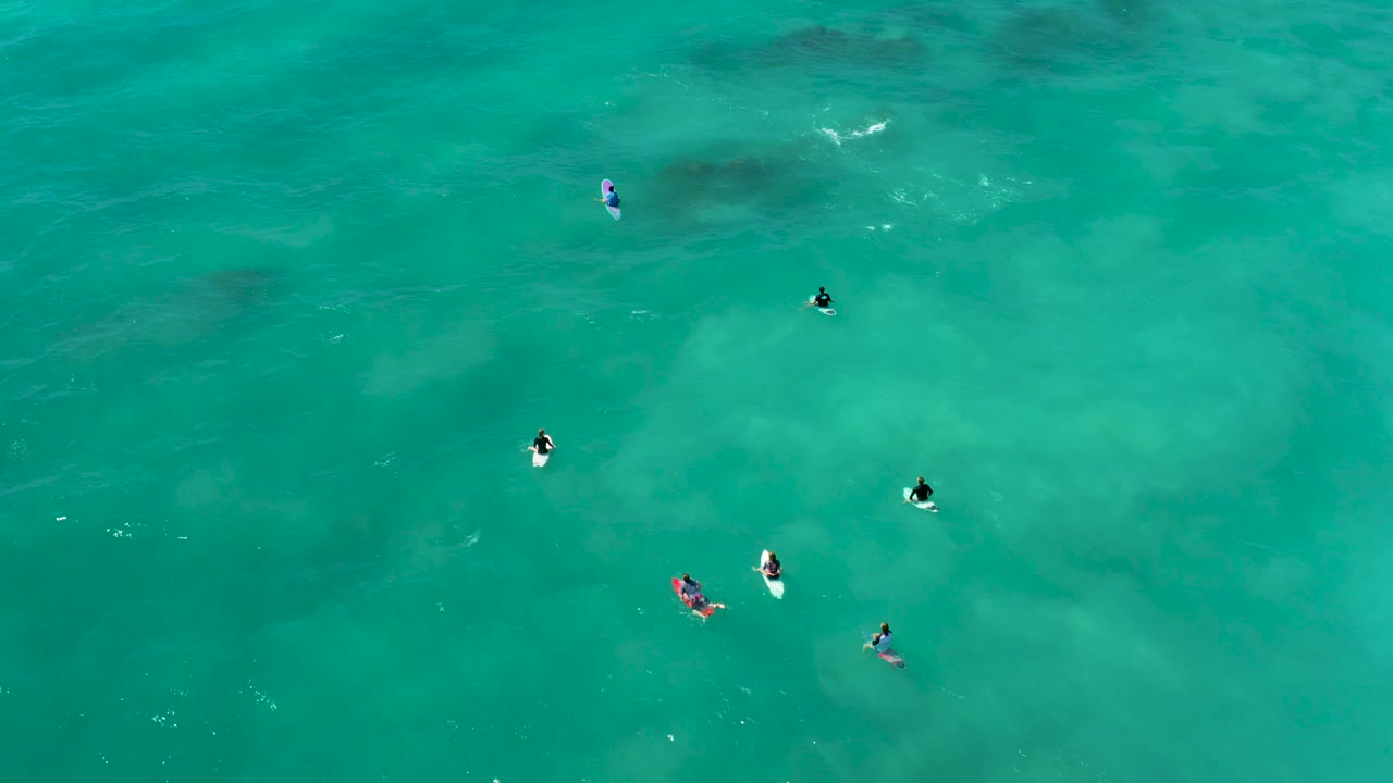 Overhead frame at Marquis Los Cabos showing several small surfers floating on boards in clear turquoise water with pale seabed patches beneath them.
