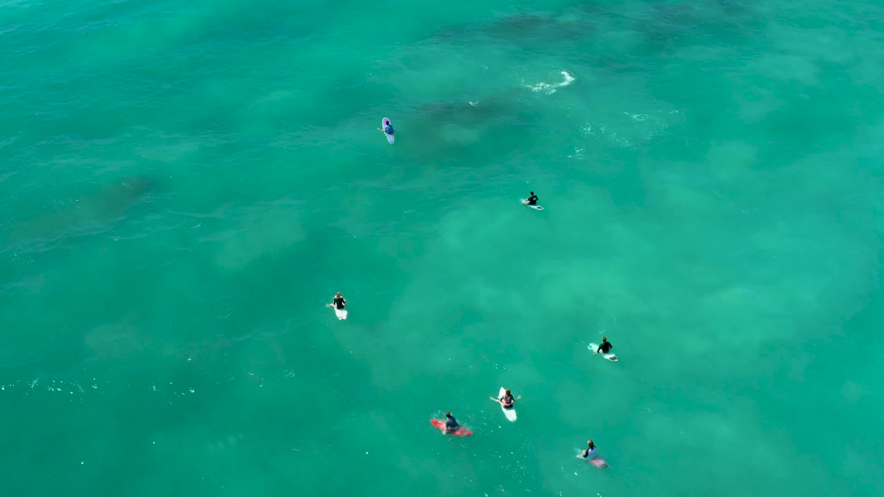 High overhead view at Marquis Los Cabos showing scattered surfers in turquoise water, with a few bright white wave streaks near the upper part of frame.