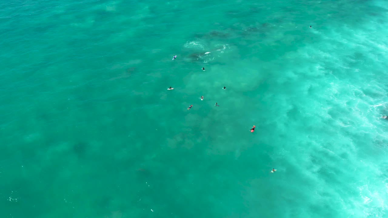 Overhead view at Marquis Los Cabos showing small swimmers scattered in clear turquoise ocean with pale wave wash near the right side.
