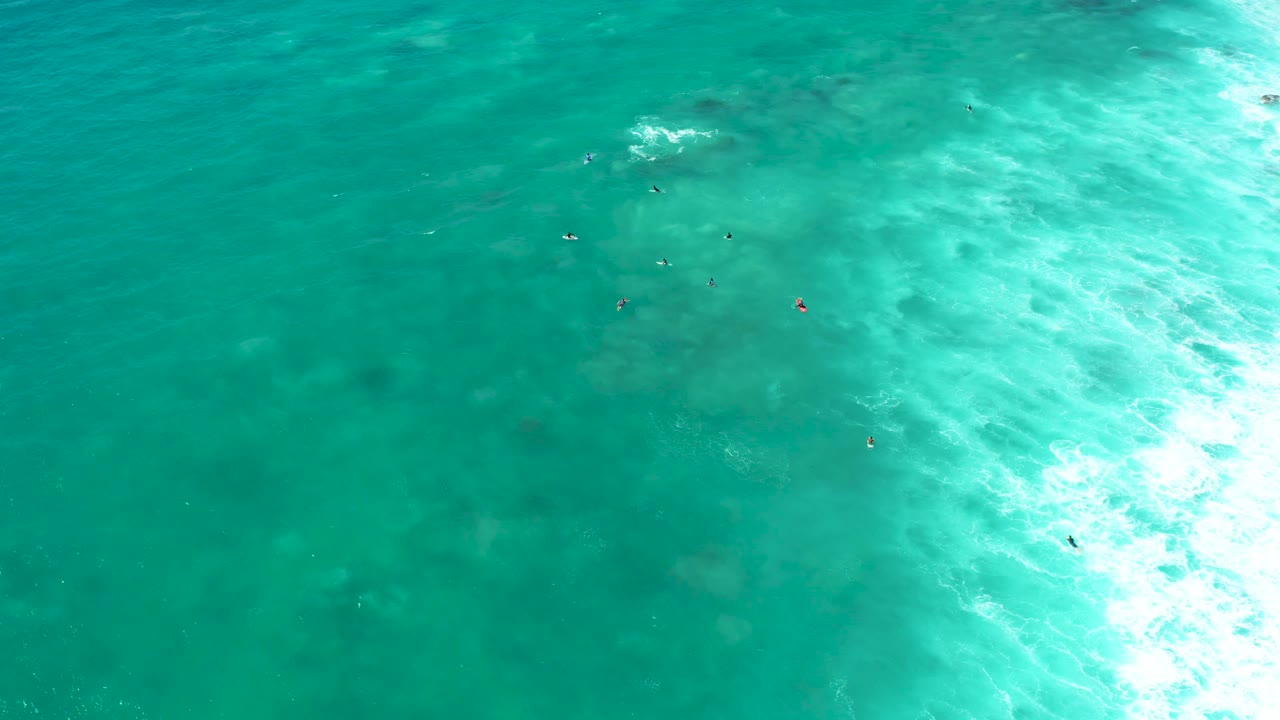 Overhead view at Marquis Los Cabos showing several small swimmers in clear turquoise water, pale seabed below, and bright foamy water along the right edge.