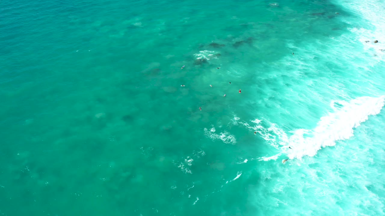 Overhead view at Marquis Los Cabos of clear turquoise ocean shallows with several small swimmers near center and bright white surf foam along the right side.