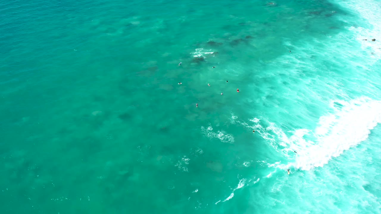 Aerial view at Marquis Los Cabos with a wide turquoise water field, tiny visible people in the shallows, and a bright foam line pressing in from the right.