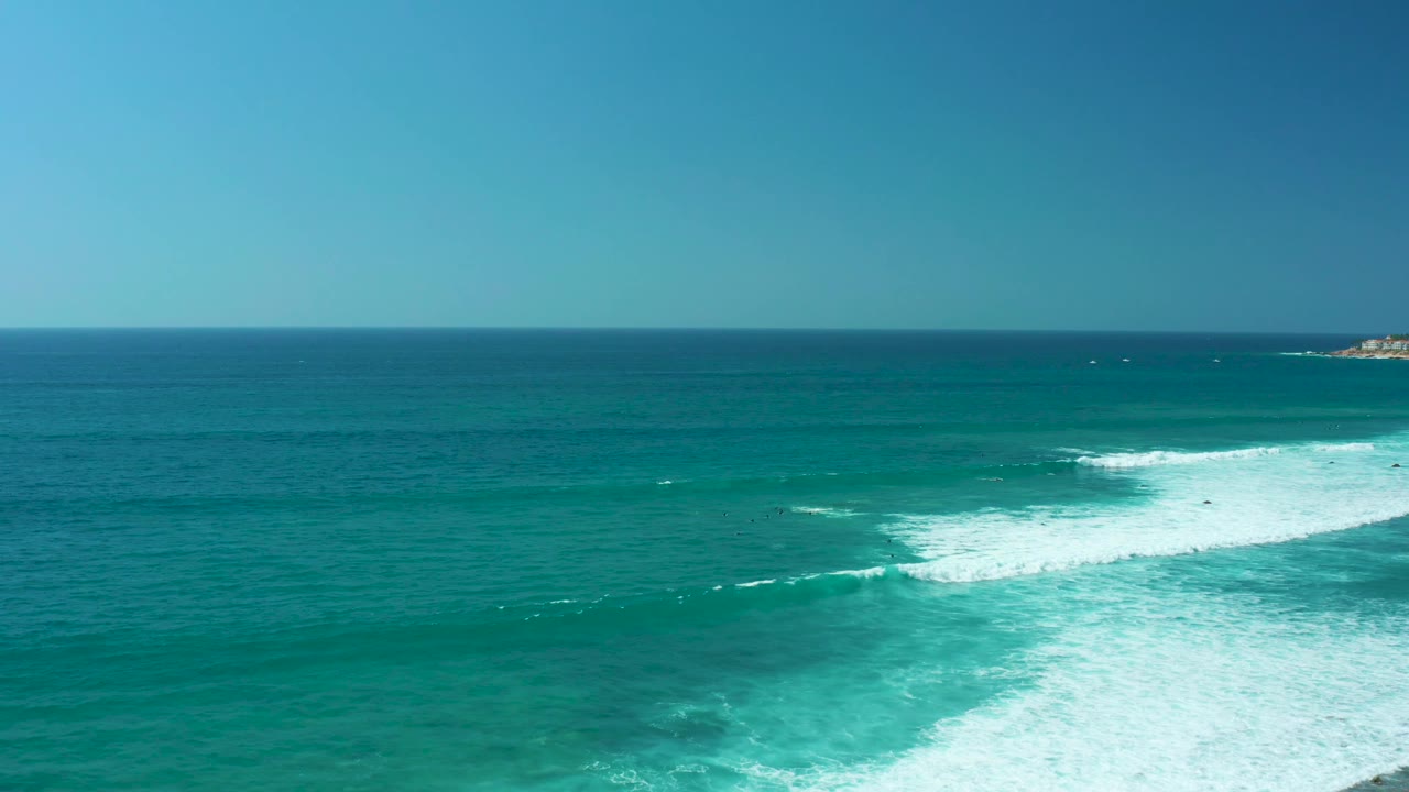 Aerial ocean frame from Marquis Los Cabos showing white foam expanding across the turquoise midground, darker blue water beyond the horizon line, and no visible people.