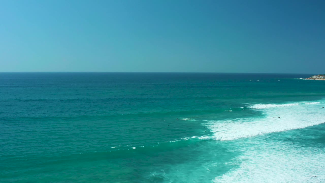 Aerial view over Marquis Los Cabos water with the breaking wave shifted farther right, open teal water across the center, a high horizon, and no visible people.