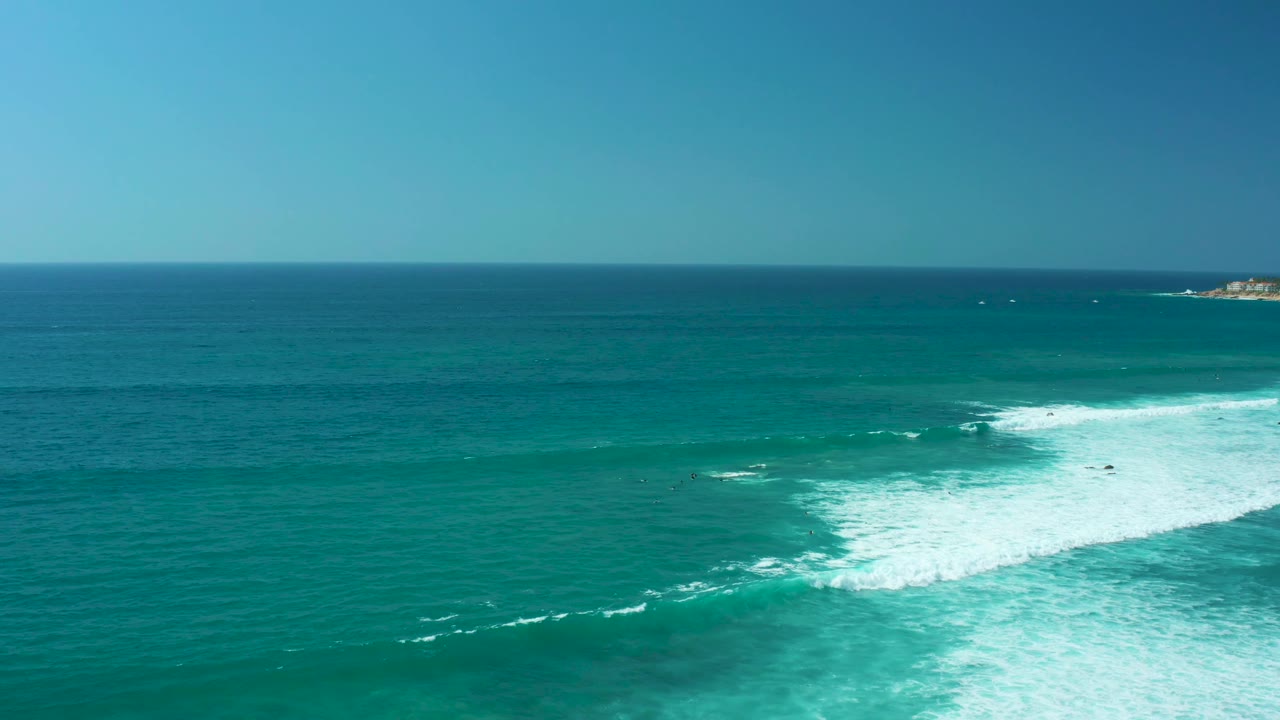 Aerial Marquis Los Cabos ocean view with white foam cutting diagonally through the lower right, teal water filling the midground, distant land near the horizon, and no visible people.
