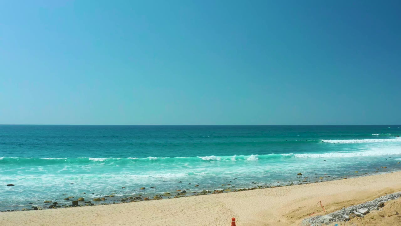 At Marquis Los Cabos, white foam spreads across blue-green water toward the sandy lower edge, rocks line the shallows, a small red object remains near the beach edge, and no visible people appear.