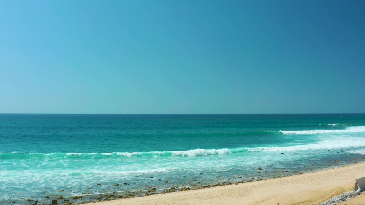 At Marquis Los Cabos, the surf line shifts right across turquoise water, pale sand appears at the lower right, shallow rocks remain visible, the sky is clear, and no visible people are present.