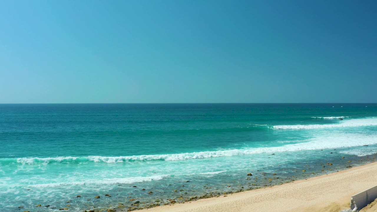 At Marquis Los Cabos, a wider band of white surf crosses the lower water, deep blue ocean sits beyond it, the sandy beach clips the lower right corner, and no visible people appear.