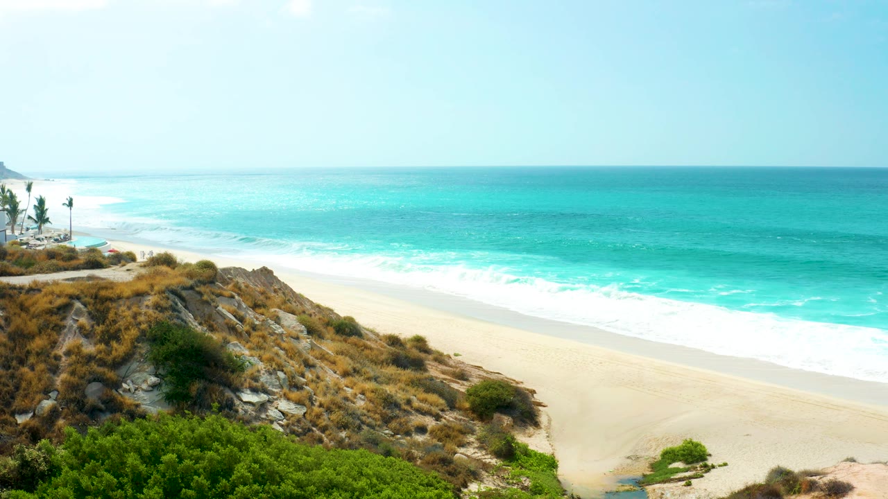 At Marquis Los Cabos in Baja California Sur, turquoise surf meets a pale sandy beach below a rocky hillside with green shrubs and no visible people.