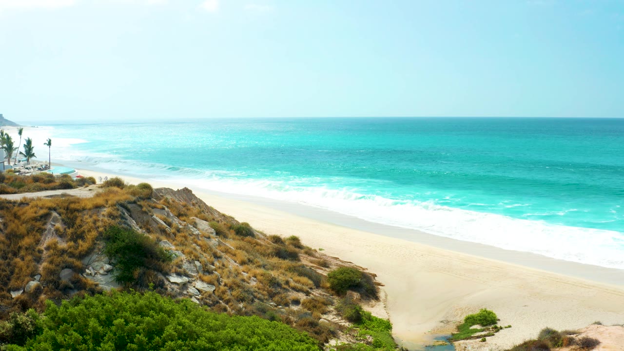 A bright view over the Marquis Los Cabos shoreline shows white waves spreading along the sand, with dry grasses and shrubs on the rocky slope and no visible people.