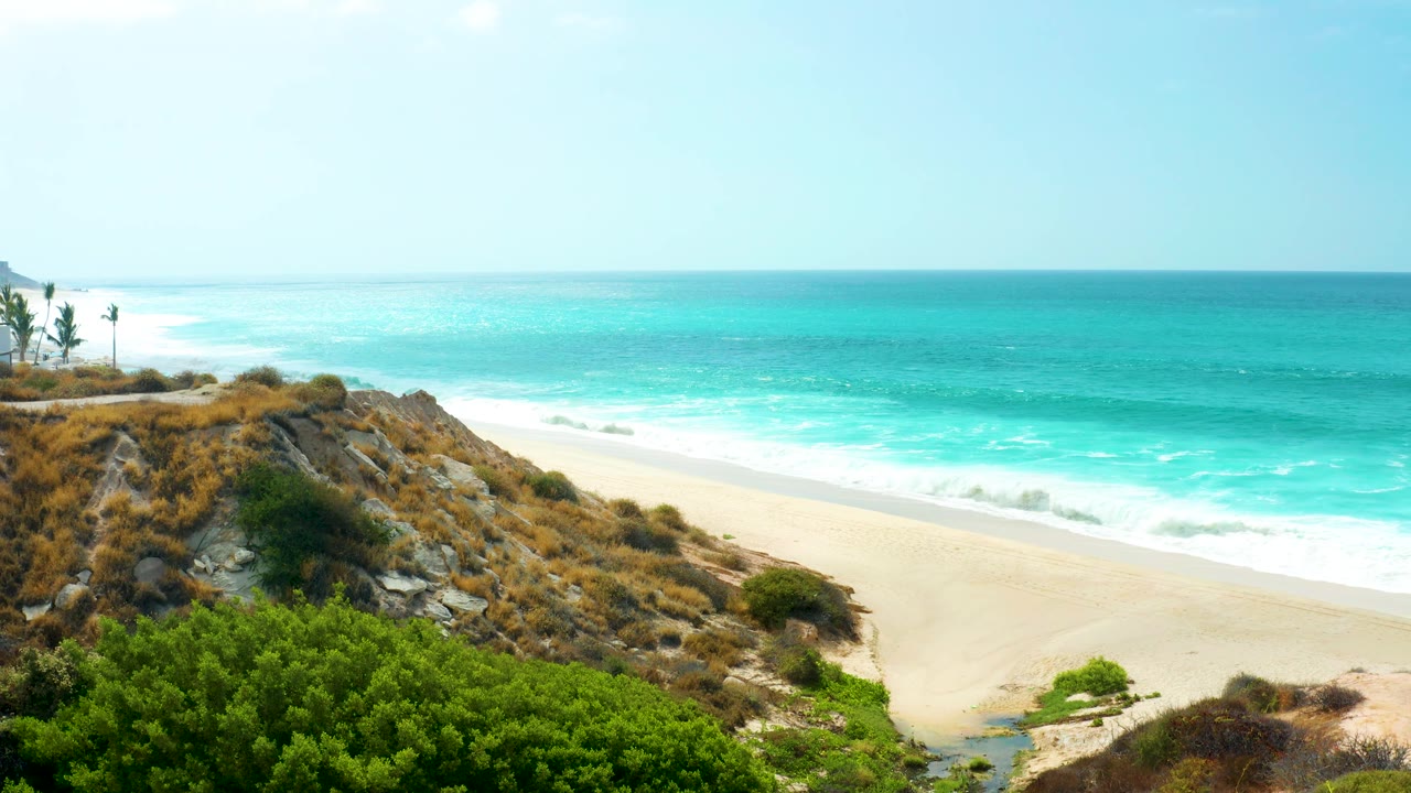 Mid-clip, the elevated view holds on turquoise water rolling toward sand below the rocky Marquis Los Cabos hillside, with shrubs in front and no visible people.