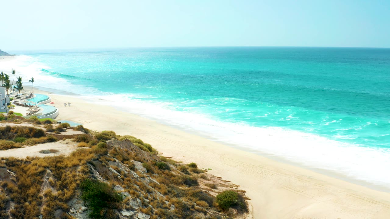 At 0:01, the Marquis Los Cabos shoreline shows a broad white foam line moving up the beach, with turquoise water beyond and scrub-covered rocks filling the lower foreground.