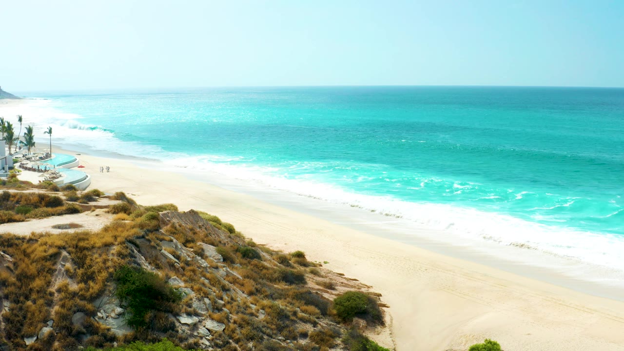 At 0:03, turquoise water and white foam run diagonally along the Marquis Los Cabos beach, while rocky scrub dominates the lower frame and palms remain visible near the left edge.