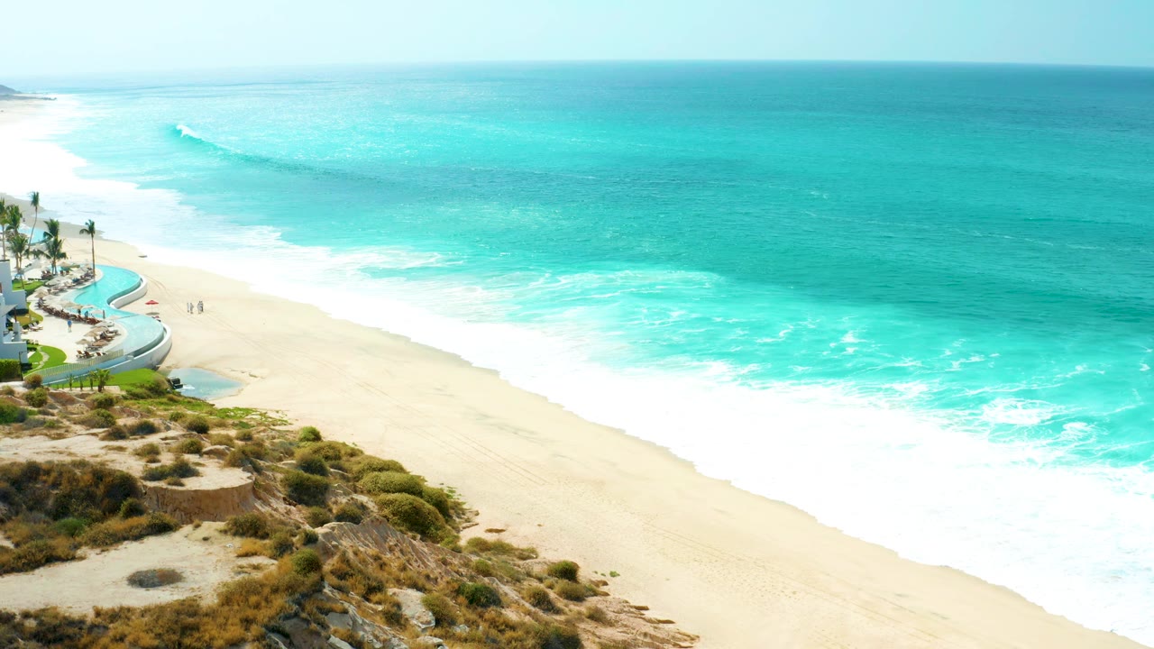 Opening frame of Marquis Los Cabos shoreline with turquoise ocean, a white breaking wave, pale sand, cliff vegetation, and resort-side pool shapes at the left edge.