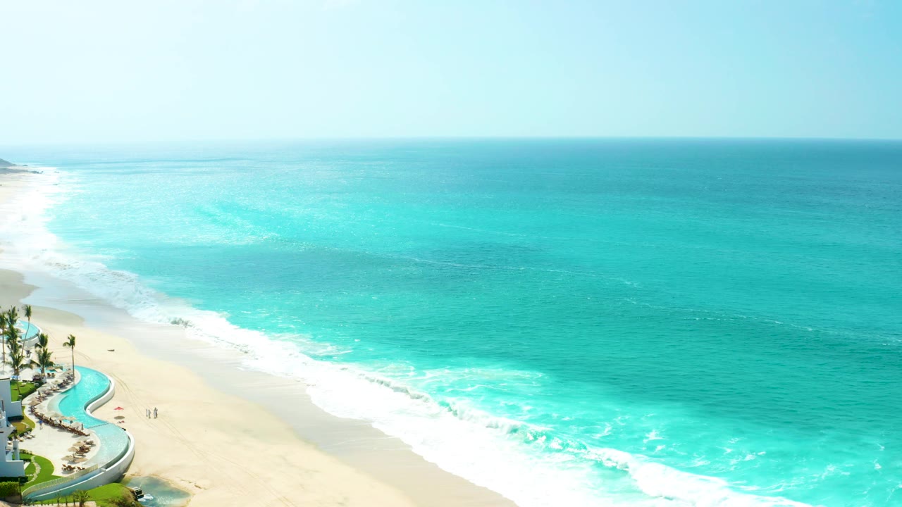 Opening aerial frame at Marquis Los Cabos, Mexico, with turquoise water, white surf, pale sand, a curved pool at the left edge, palms, and tiny distant beachgoers on the sand.