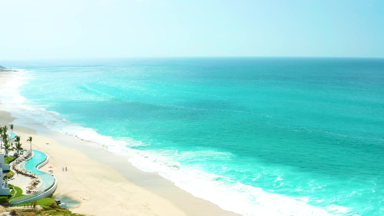A brighter aerial frame of the Marquis Los Cabos beach, with broad turquoise water across the top, white wave bands along the shore, and the resort pool still visible at lower left.