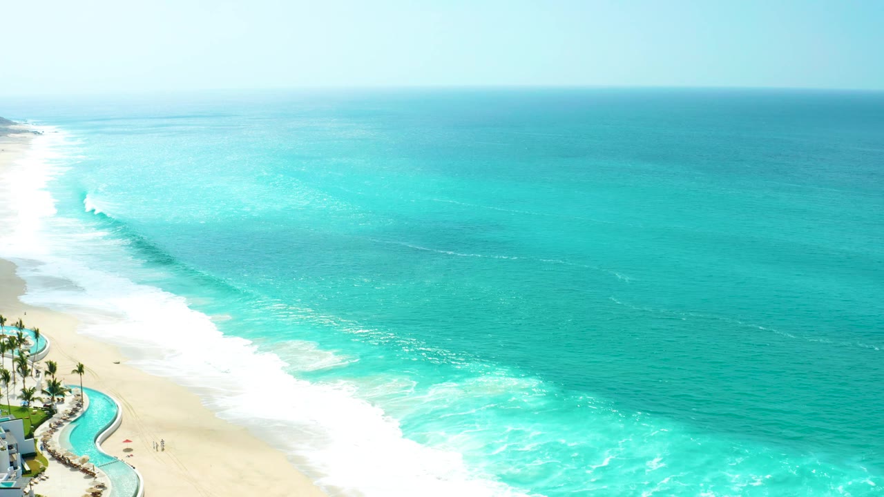At 0:01, the Marquis Los Cabos shoreline shows a broad white foam band across the lower frame, turquoise water beyond it, palm trees at left, and no visible people.