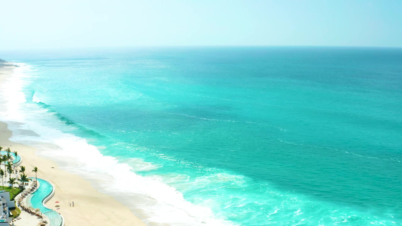 At Marquis Los Cabos, a breaking wave angles in from the left toward the sand, with open turquoise sea across the frame and no visible people.