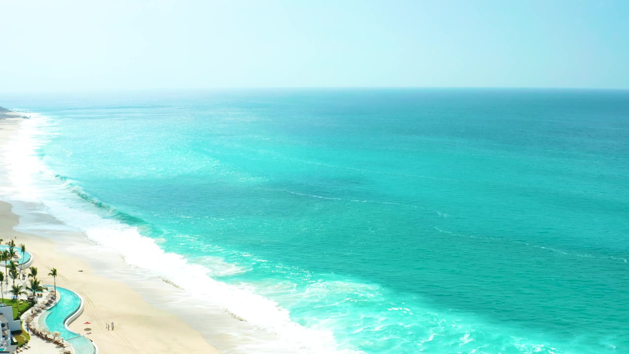 Mid-clip view of Marquis Los Cabos beach with bright turquoise water, a long foamy edge on the sand, resort palms and pool at the lower left, and no visible people.