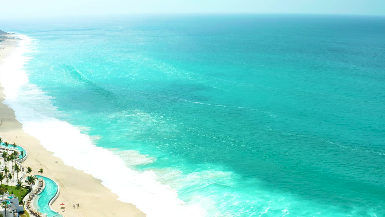 Large turquoise wave forming offshore beside a sandy beach and pool area.