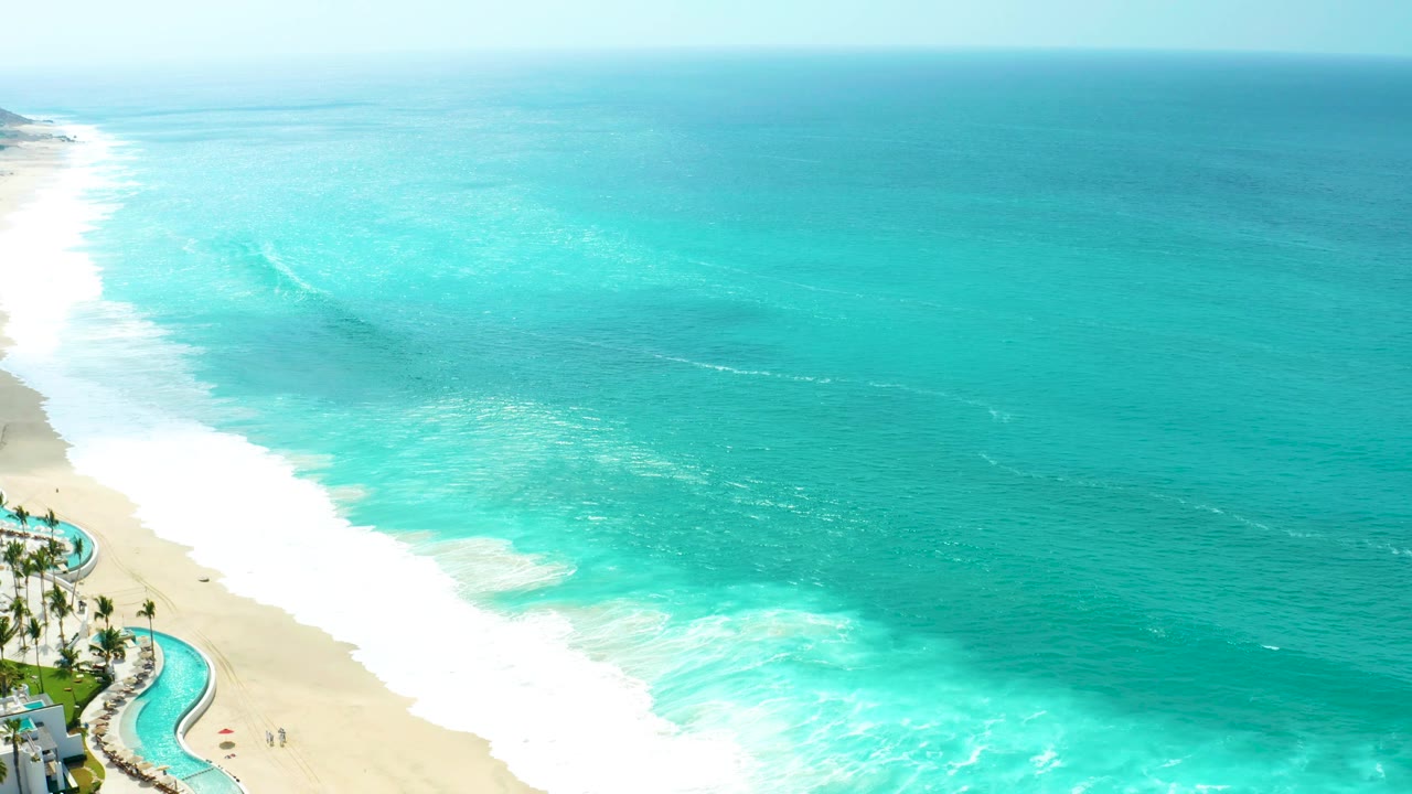 The Marquis Los Cabos shoreline stays in the lower left as white surf pushes diagonally across the sand beneath a broad field of turquoise ocean.
