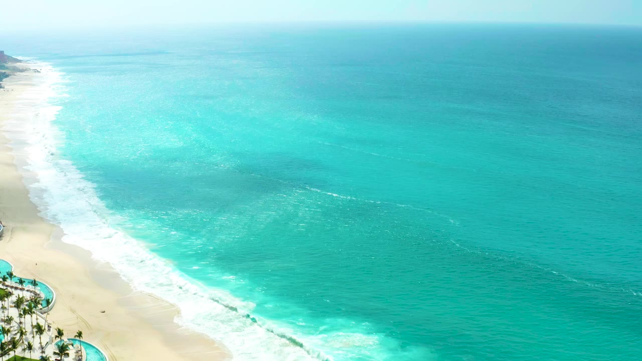 The second frame shows a long white foam band sliding along the Marquis Los Cabos shoreline, with pale sand on the left and open turquoise ocean to the right, with no visible people.