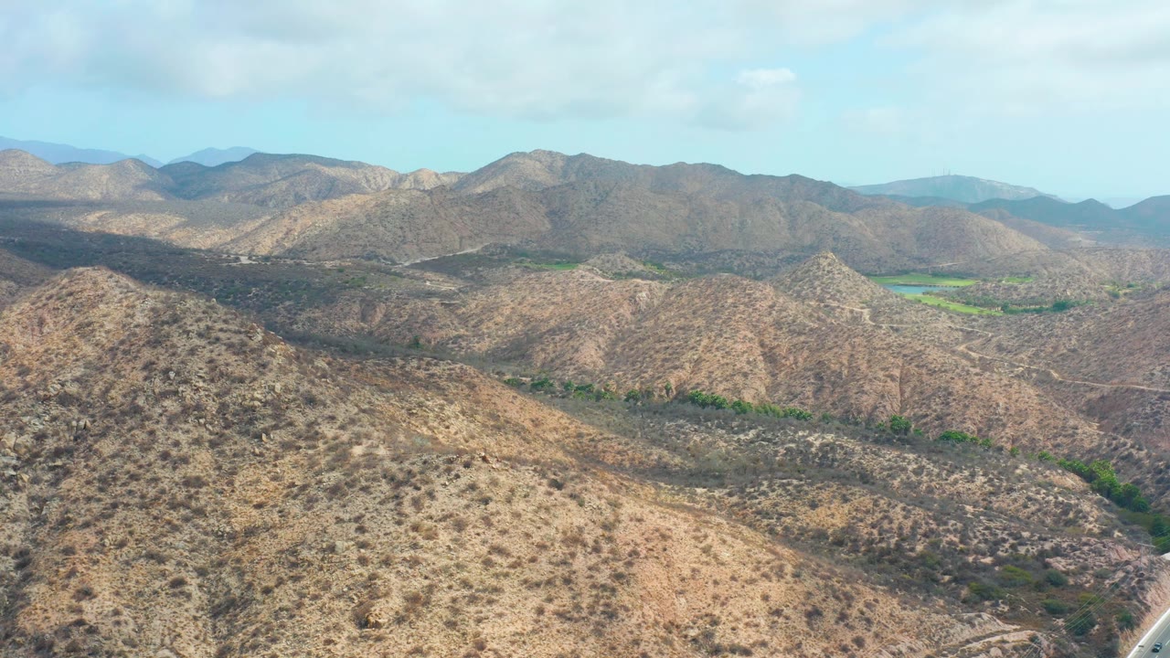 Second frame from Marquis Los Cabos shows the foreground ridge slightly more centered, with brown scrub-covered slopes, darker valley channels, green patches near the horizon, and no visible people.
